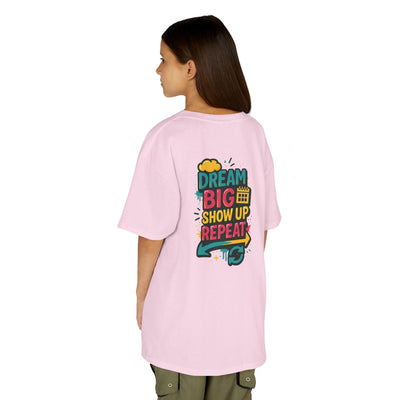 Back view of a girl in a pink motivational tee featuring 'Dream Big. Show Up. Repeat.' message, perfect for kids.