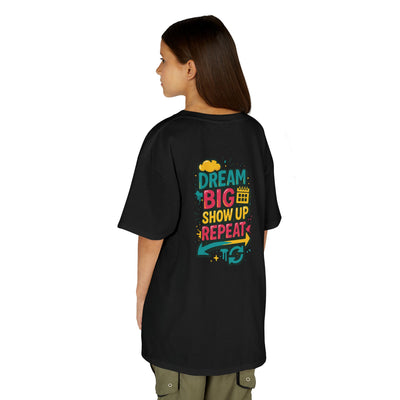 Back view of a kid wearing a black motivational tee with the phrase 'Dream Big. Show Up. Repeat.' in colorful print.