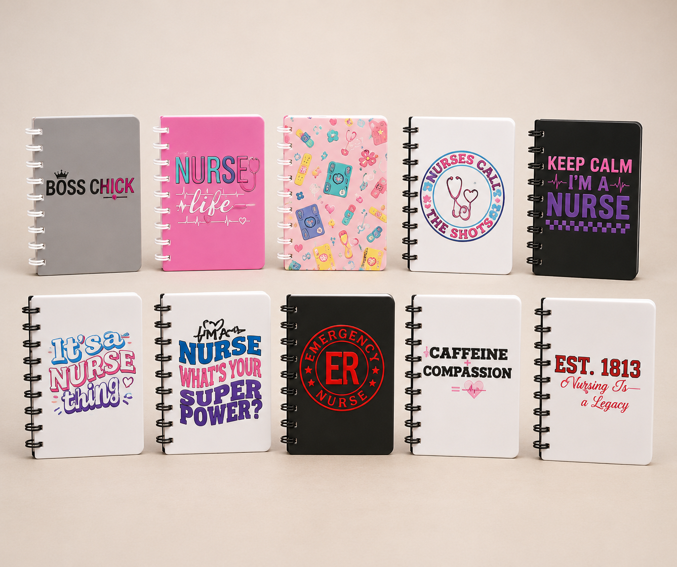 Eight spiral-bound notebooks arranged in two rows on a light beige background. The notebooks feature various colorful and monochrome designs with nurse-themed quotes and graphics, including 'BOSS CHICK', 'NURSE like', 'NURSES CAN'T BE SHOT!', 'KEEP CALM I'M A NURSE', 'It's a NURSE thing!', 'NURSE WHAT'S YOUR SUPER POWER?', 'EMERGENCY ROOM NURSE', and 'CAFFEINE COMPASSION' and 'EST.